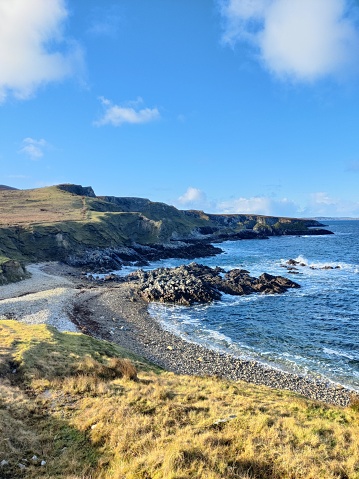 Coastal bliss: Waves meet shore, cliffs stand tall under a blue sky, framed by reedy grass in the foreground