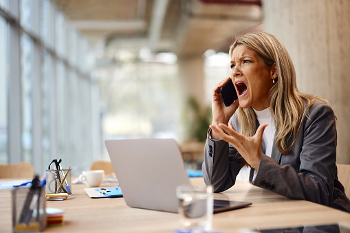 Displeased female entrepreneur screaming at someone over cell phone while working on laptop in the office.