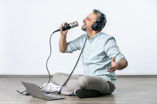 Handsome man with headphones and microphone connected to laptop sitting on the floor against white background. E-learning singing lessons concept.