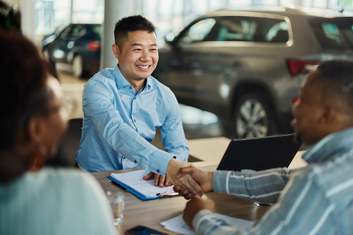 Happy Asian car salesman came to a successful agreement with black couple during a meeting in a showroom.