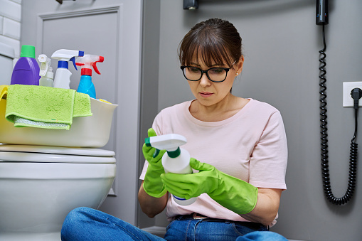 Woman reading label on spray cleaner, composition is chemical, organic, instructions for use, in bathroom. Household chemicals for cleaning house. Housekeeping, housework, housecleaning, cleaning service concept