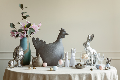 Warm and spring dining room interior with easter accessories, round table, vase with green leaves, cake, colorful eggs, rabbit sculpture and personal accessories. Home decor. Template.