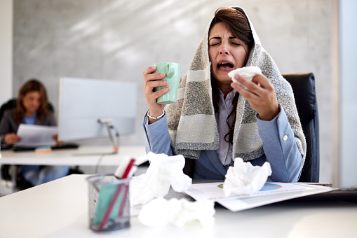 Displeased businesswoman crying in despair while being sick in the office. Her colleague is in the background. Copy space.