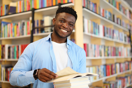 A diligent young African student in a university library, studying amidst bookshelves, preparing for exams with a smile.