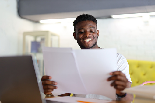 An African American businessman sits at his desk, smiling as he reads through paperwork, embodying professionalism and dedication in his work.