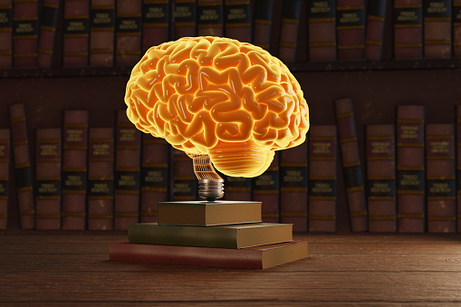 Glowing yellow human brain acting like a light bulb on a stack of hard cover books with a bookshelf as background. Illustration of the concept of knowledge, education and studying