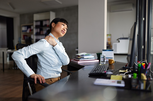 Japanese businesswoman holding her back in pain while working on desktop PC at corporate office. Copy space.