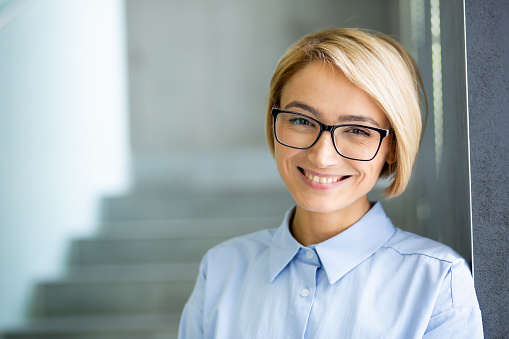 Close-up portrait of young beautiful blonde business woman in glasses, female worker at workplace inside office building smiling looking at camera.