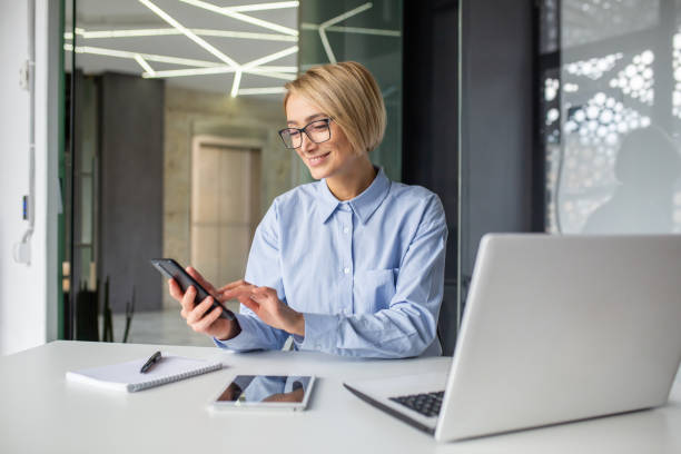 istock Careless attractive office worker using cellular phone while turning away from laptop in workplace. Inattentive blonde woman checking social media and mails instead of finishing business project 2032460776