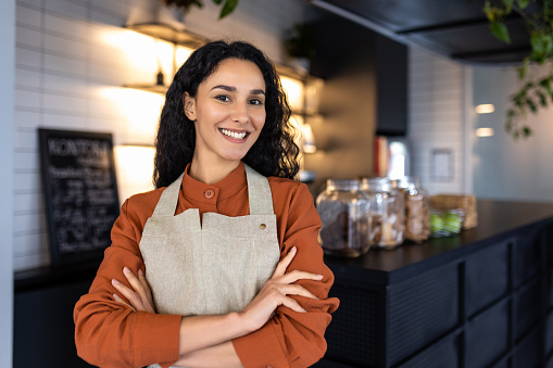 Portrait of young joyful successful Latin American small business owner, slim boss smiling and looking at camera with crossed arms, businesswoman at entrance to cafe restaurant, inviting visitors.