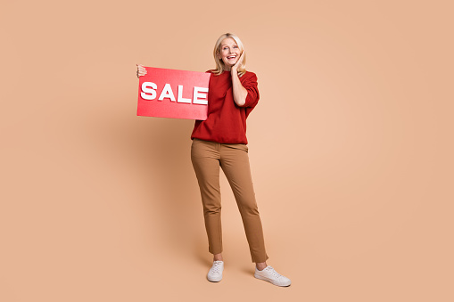 Full length photo of excited funky lady dressed red pullover hand arm cheek holding sale card isolated beige color background.