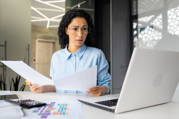 istock Thinking upset business woman holds contract papers and documents in hands, financier compares and analyzes performance indicators, works inside office looks at laptop screen 2032491430
