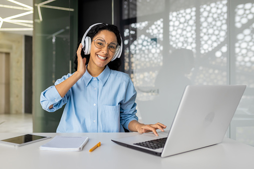 Portrait of young beautiful woman at workplace inside office, businesswoman programmer in headphones smiling and looking at camera, female worker listening to music and working with laptop.