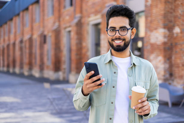 istock Relaxed middle eastern man holding telephone and plastic coffee cup while walking during daytime. Positive brunet freelancer with flexible work schedule taking break for beverage and scrolling feed 2032515598