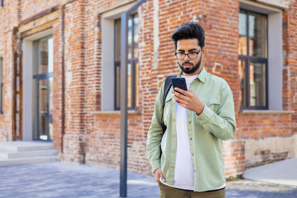 istock Serious young Indian man standing with backpack on city street, using mobile phone, reading and writing messages, making phone calls 2032515600