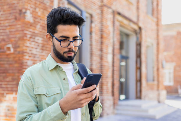 istock Young indian guy in eyeglasses looking at smartphone screen while standing with backpack outdoors. Focused young man checking online classes schedule for attending courses in university campus 2032515623