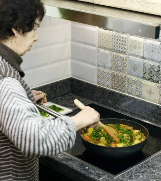 An elderly woman prepares a traditional dish of green beans, carrots, and potatoes in her kitchen