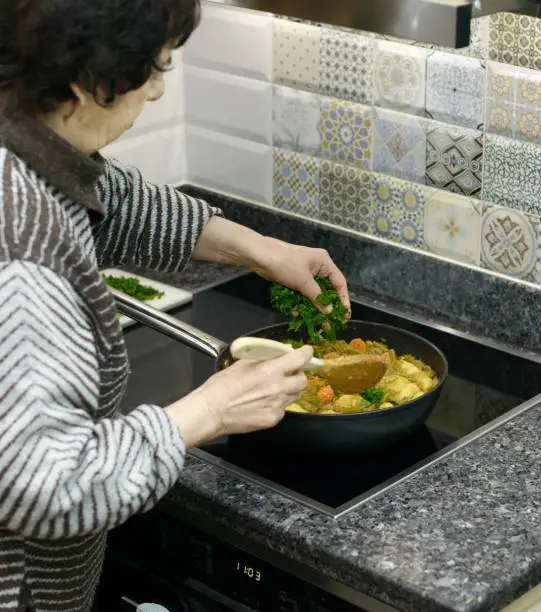 An elderly woman is sprinkling herbs onto a pan of sautéed vegetables on a stove
