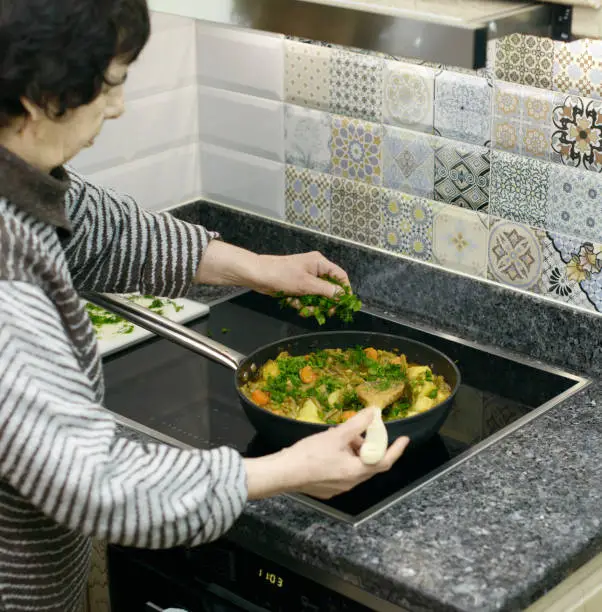 A senior woman carefully seasons a pan of simmering vegetables with fresh greens