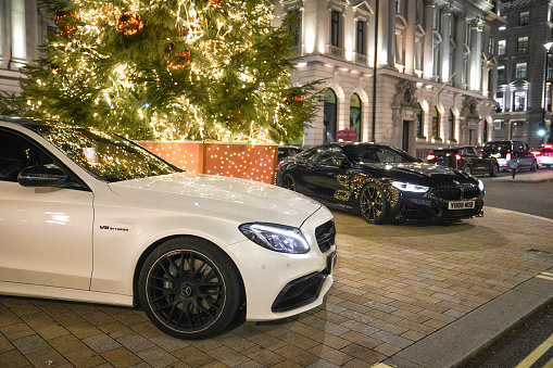 Waterloo Place on Regent Street St. James, City of Westminster, London, UK - 11th November 2023: A stylish black BMW parked beside a festive Outdoor Christmas tree in Waterloo Place, Regent Street St. James, City of Westminster, London, UK. The night street view captures the elegant ambience of this seasonal scene. Christmas festive tree in Waterloo Place near London - Pall Mall at night.