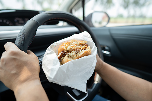 Asian lady holding hamburger and French fries to eat in car, dangerous and risk an accident.