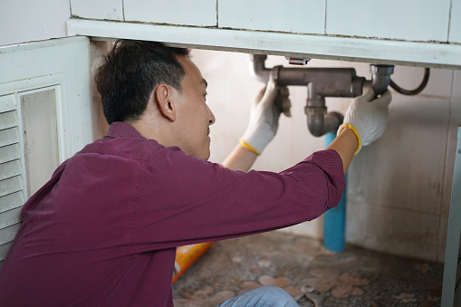 A male plumber repairs a pipeline or drain under the sink in kitchen room.