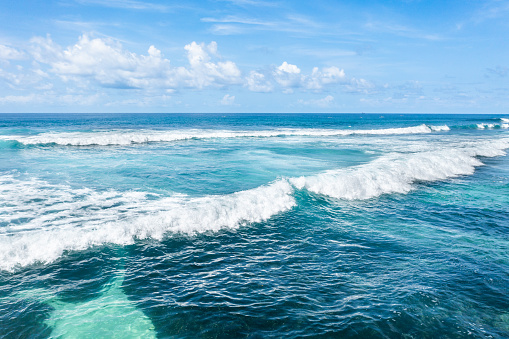 Aerial view of turquoise ocean waves under a blue sky scattered with white clouds. The crystal clear water reveals different shades of blue and green as the waves gently crash, creating white foam.