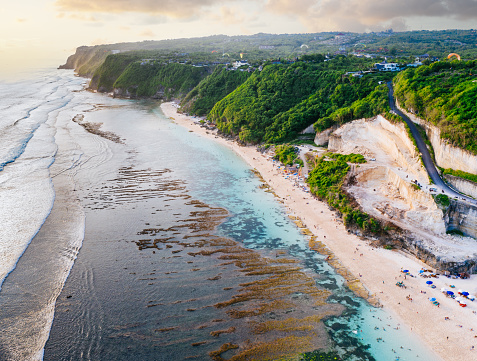 An aerial view reveals a coastal landscape bathed in the warm hues of sunset. Turquoise waters gently lap against sandy beaches, while lush greenery blankets the elevated terrain. A winding path or road winds through the verdant landscape, leading toward a small town nestled in the distance. The serene beauty of this scene invites viewers to immerse themselves in the idyllic coastal paradise. Melasti beach, Bali.