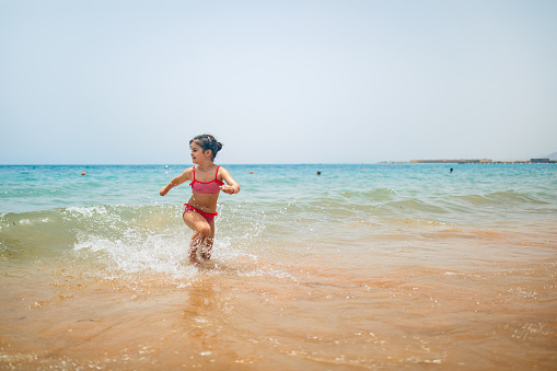 Rear view of a little girl running on the beach in a swimsuit and a straw hat in hand. Full body shot of smilling girl in blue swimsuit, enjoying sandy beach and crystalline sea of Mnemba beach in Zanzibar, with copy space. Concept of beach summer vacation with kids.