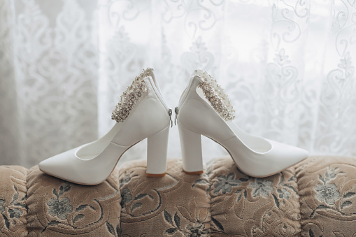 Photo of details at the wedding. White chic shoes with a decoration on the foot, standing on the sofa, near the window. Contour line. Shadows and light.