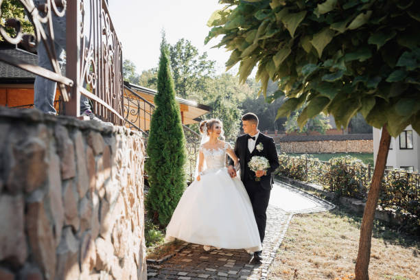 istock A groom in a black suit and a blonde bride stand embracing by a stone wall under a tree. 2032622190