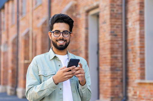 Casual arab male looking at camera with wide smile while using cellphone on brick building background. Careless gadget user in classic spectacles enjoying playing new online mobile application.