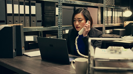 Law officer verifying details for the private investigation, working in archive room. Detective cross checks information on landline phone call, contacting people from records. Camera B.