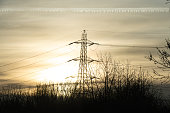 A distant electrical pilon, pylon with winter bushes without their leaves in the foreground and a vivid bright sky with the sun setting and a vapour trail running along the top of the sky