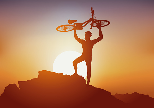 Concept, with a cyclist who, as a sign of victory, lifts his mountain bike to the top of a mountain in front of a sunset.