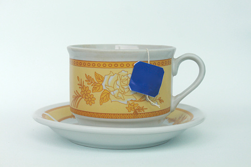 An empty white ceramic cup with a luxurious floral pattern with a tea bag on a blue label. Isolated in white background.