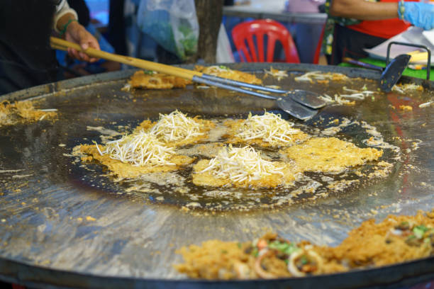 Thai street food chef preparing a traditional crispy oyster omelette, garnished with fresh bean sprouts on a large iron griddle