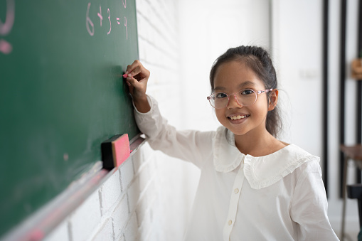 An Ethnic teacher is leading a class of elementary school children. There are various posters on the wall, and drawings on the chalkboard. Students are putting up their hands to answer a question.