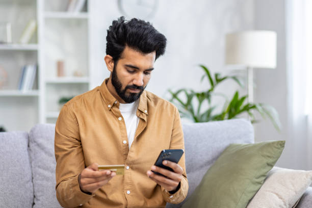 istock Indian young man sitting on sofa at home and using mobile phone and credit card 2032762872