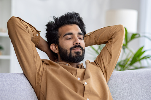 Relaxed bearded man with closed eyes resting on a couch, hands behind head in a cozy room, concept of peace and leisure time at home.