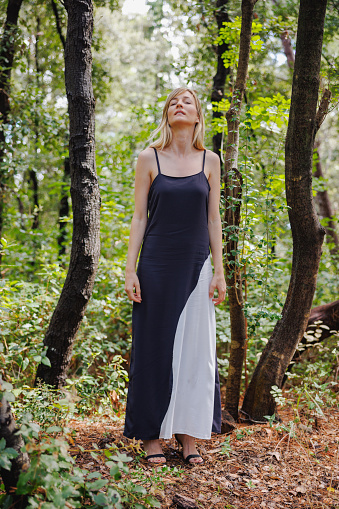 Beautiful blond woman in sleeveless dress standing on dry leaves on the ground in the forest, enjoying nature and fresh air