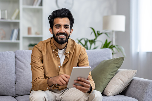 Portrait of a smiling young Indian man sitting on a sofa, holding a tablet and looking at the camera.