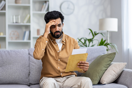 A Hindu man is holding his head with his hand in embarrassment and opening an envelope with a letter, shocked by bad news.