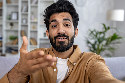 Close-up photo of a smiling young Indian man at home and talking on a video call on the phone to the camera, gesturing with his hands.