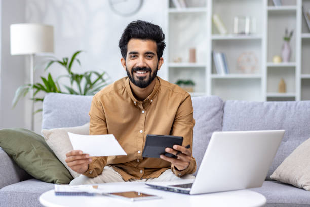 smiling young man with beard working from home, using laptop and calculator, financial planning concept - multi tasking asian and indian ethnicities asian ethnicity lifestyles imagens e fotografias de stock