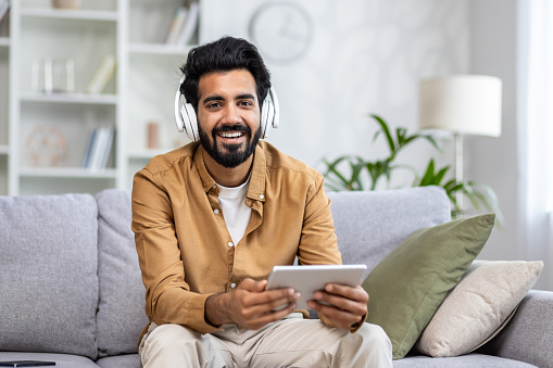 Portrait of a young Indian man wearing headphones sitting on the couch at home and holding a tablet, smiling and looking at the camera.