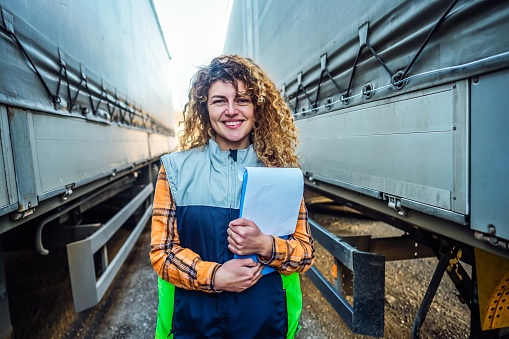 Portrait of a smiling female truck driver with shipment list standing between truck vehicles. Transportation service
