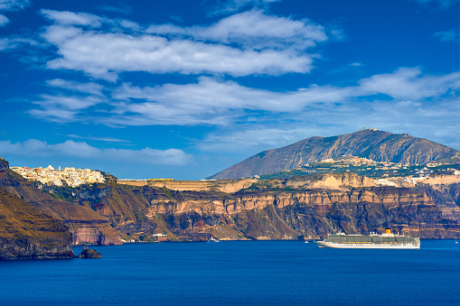 Panorama of Santorini island, Greece. Rough cliffs of volcanic island, sunny day, caldera bay, blue sky and clouds, Fira or Thira town on hilltop, cruise ship anchored in harbor. Summer cruise, vacations, travel destination, tourist attraction, sea