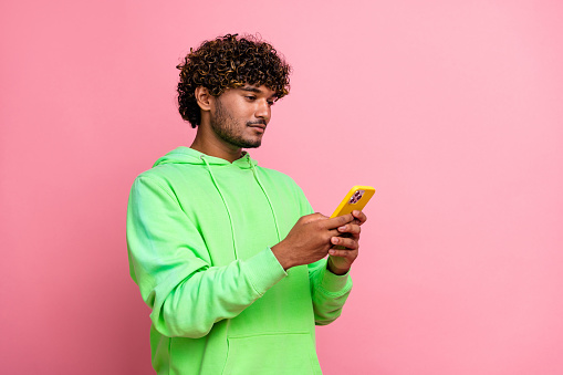 Photo of confident serious young arabian man wearing green hoodie using mobile phone for commerce isolated on pink color background.