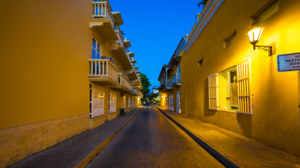 This evocative photograph captures the essence of Cartagena de Indias, Colombia, as dusk descends upon the city. The golden hues of the streetlamps cast a warm glow against the rich mustard-colored walls of the colonial buildings lining the cobblestone street. A sign indicating \